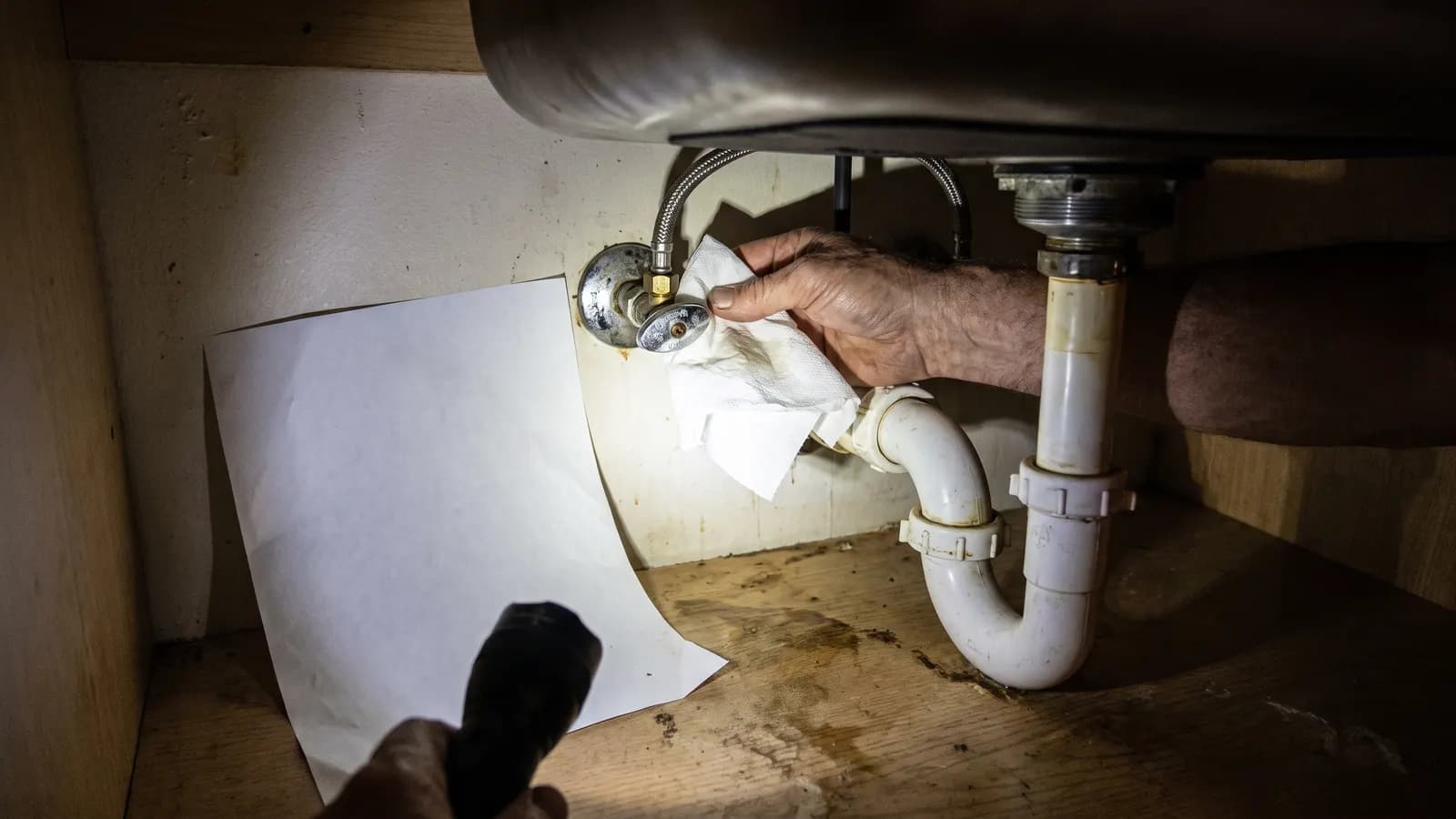 Paper towel and flashlight checking under-sink supply lines for leaks